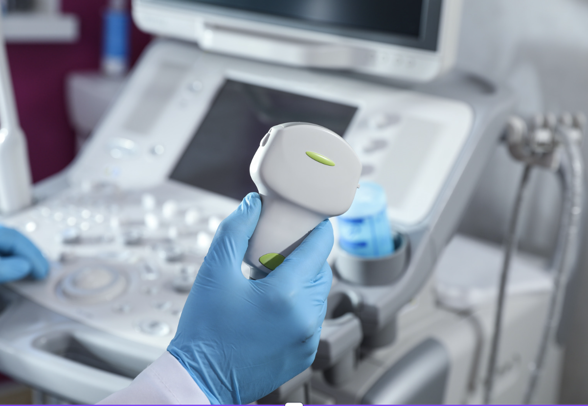 Gloved clinician holding an ultrasound probe in front of an ultrasound machine in a clinic room.